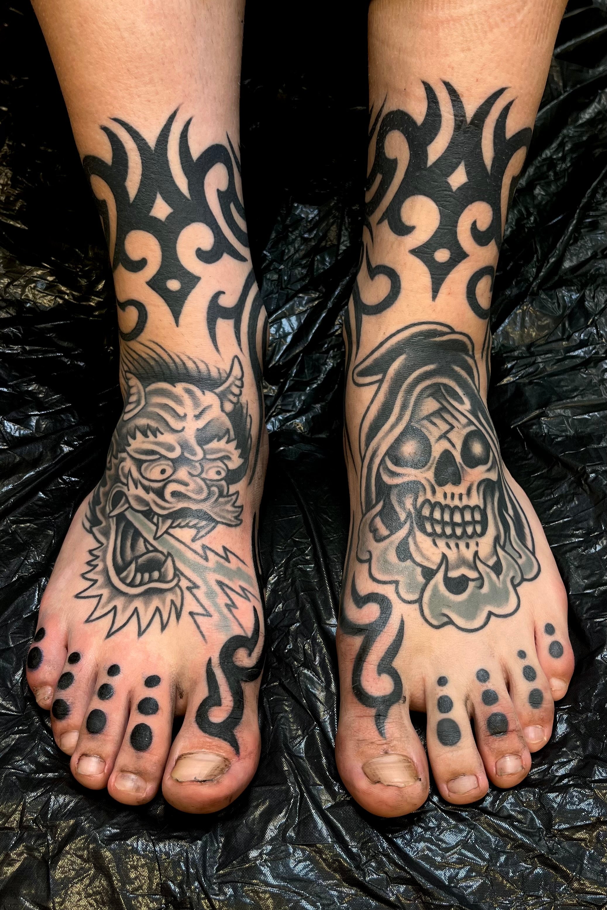 Pair of fully tattooed feet in black and grey American traditional of grim reaper skull head and an ed hardy Japanese demon head tattoo with lightening, and blackwork tribal ankle cuffs and fillers . Tattooed in London