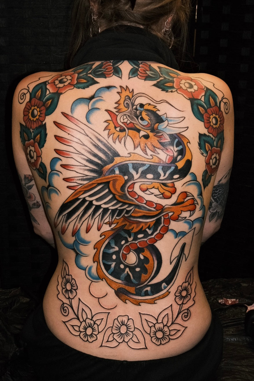 full colour american traditional backpiece of a winged dragon with a frame of flowers. 