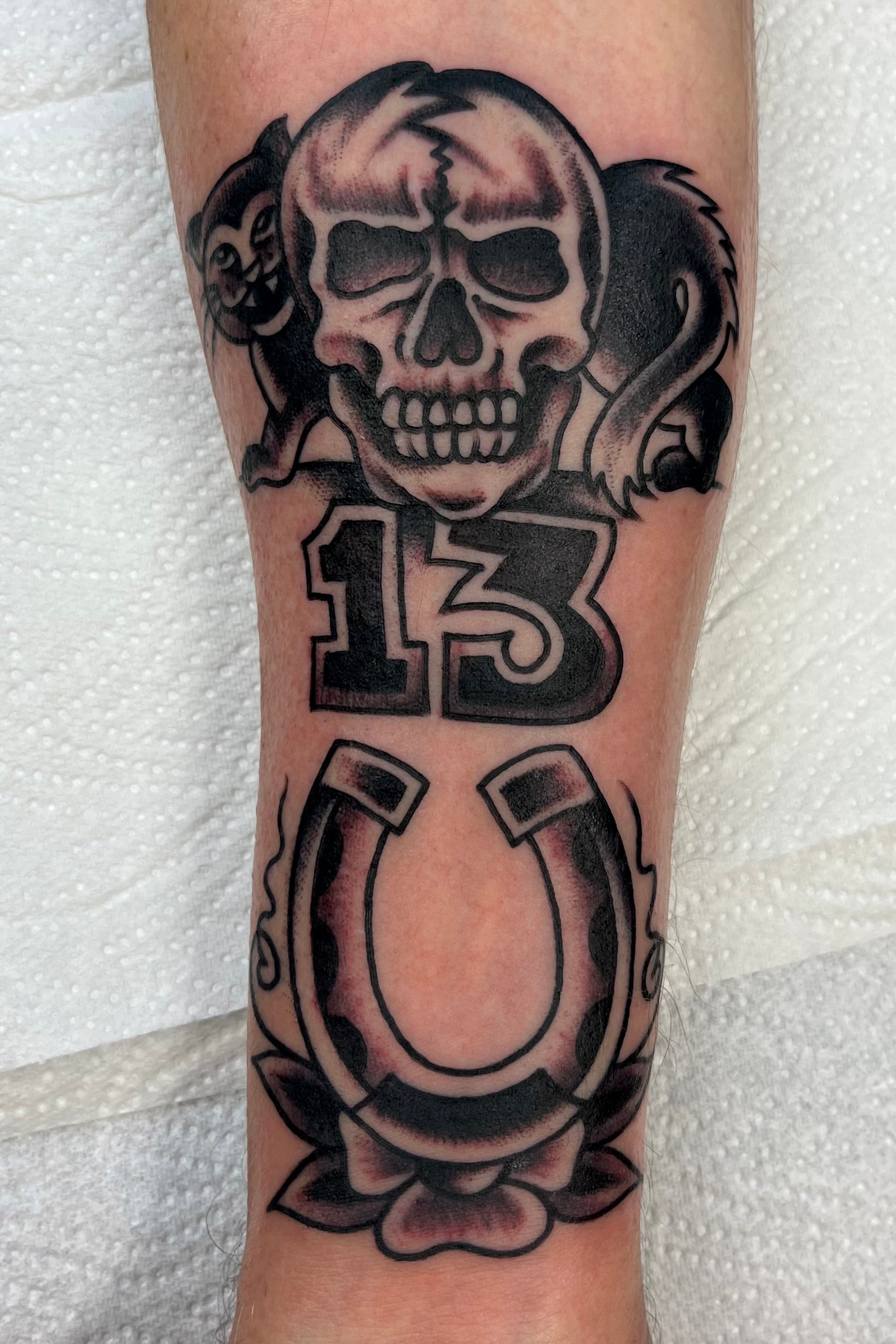 a bold american traditional black and grey tattoo of a lucky 13 black cat and a skull, with a horseshoe and flowers. tattooed in london at frith street tattoo