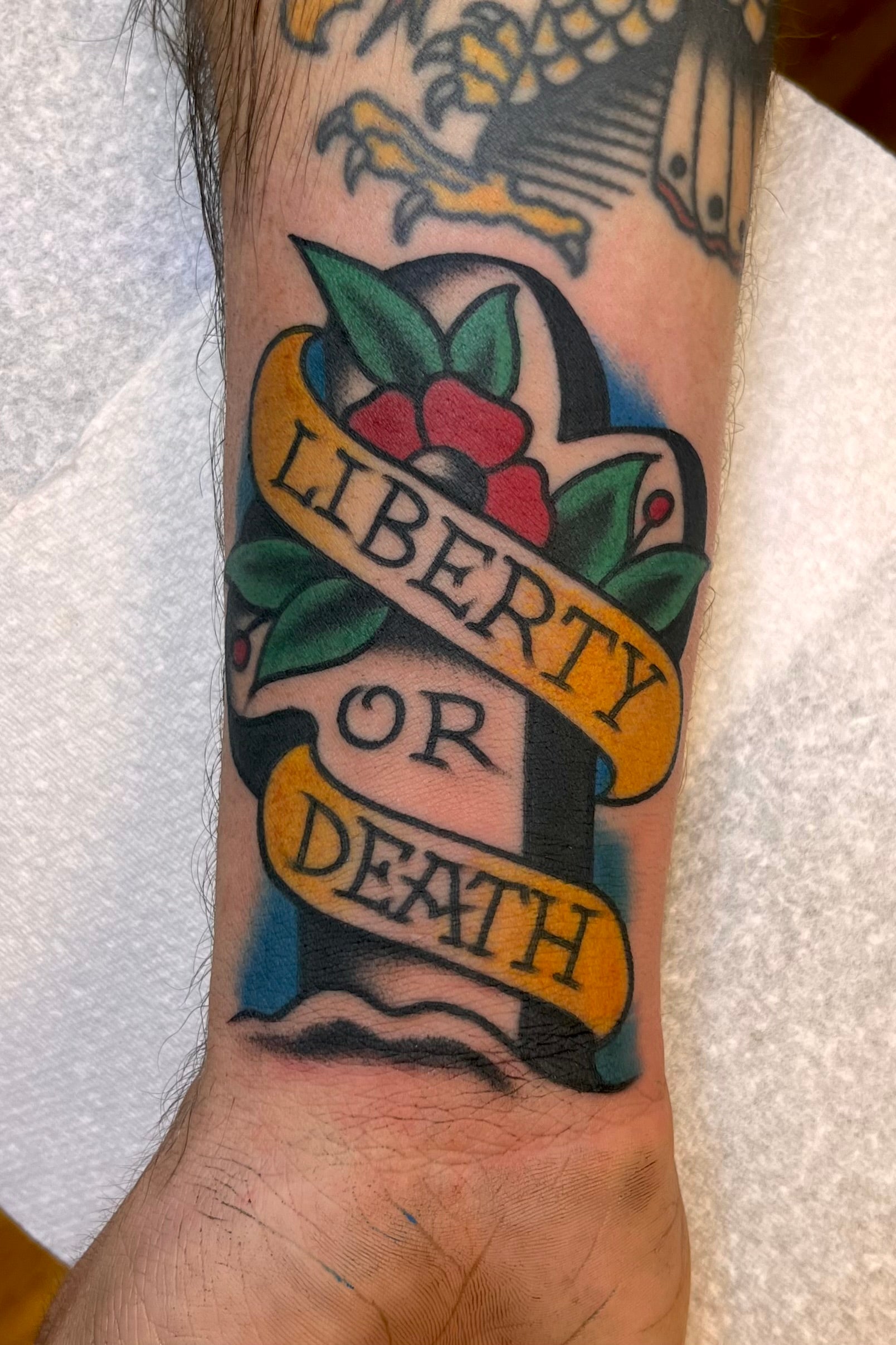 Bold American traditional classic Memorial cross tattoo with flowers and leaves, and a banner saying Liberty or death in colour. Done in Central London