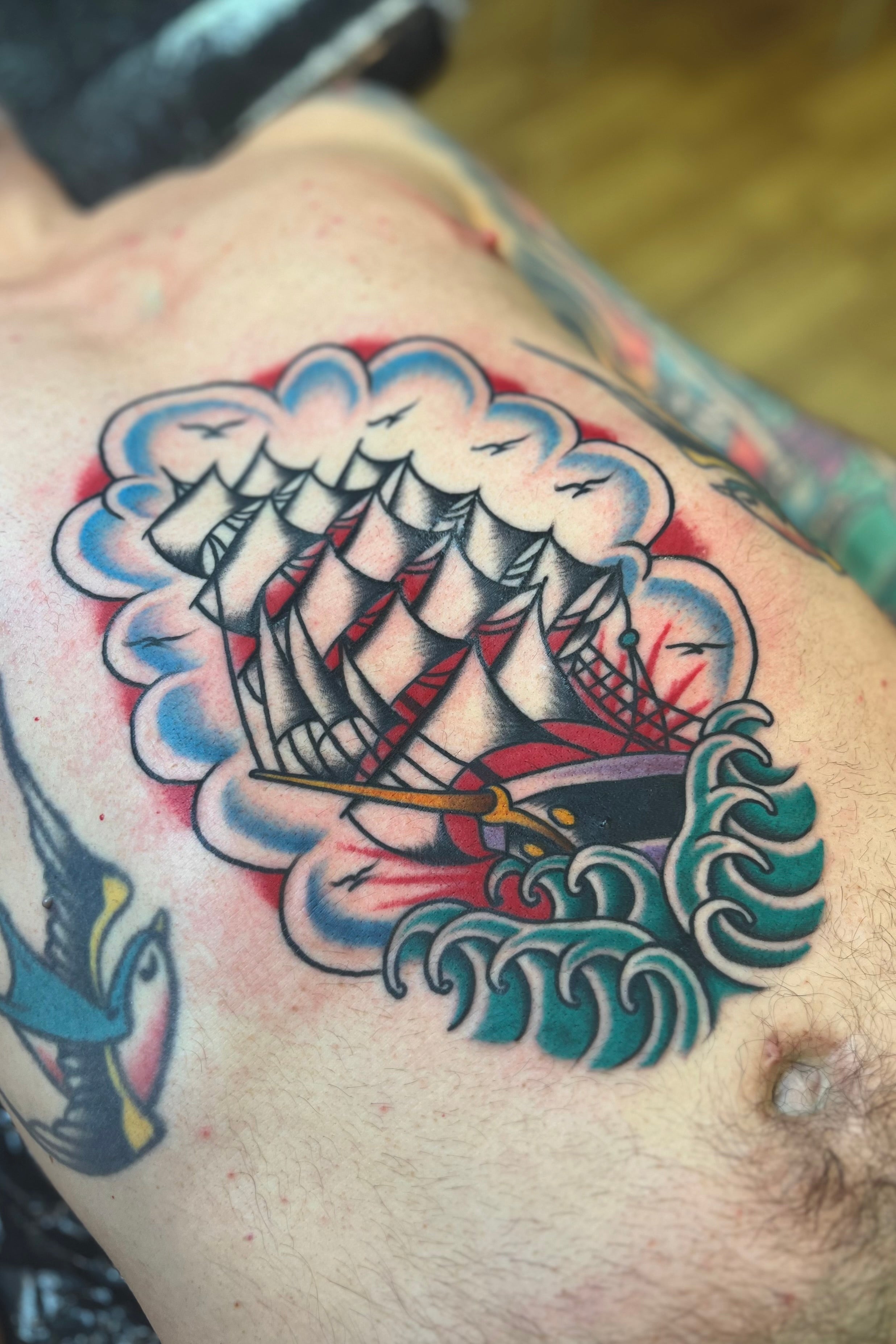 Bold American traditional classic ship front piece tattoo in bright colours with Japanese waves in the style of Bob roberts. Tattooed in London